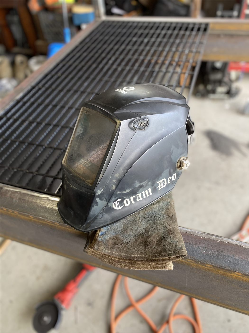 Welding helmet hand-lettered with Coram Deo, resting on steel in the shop